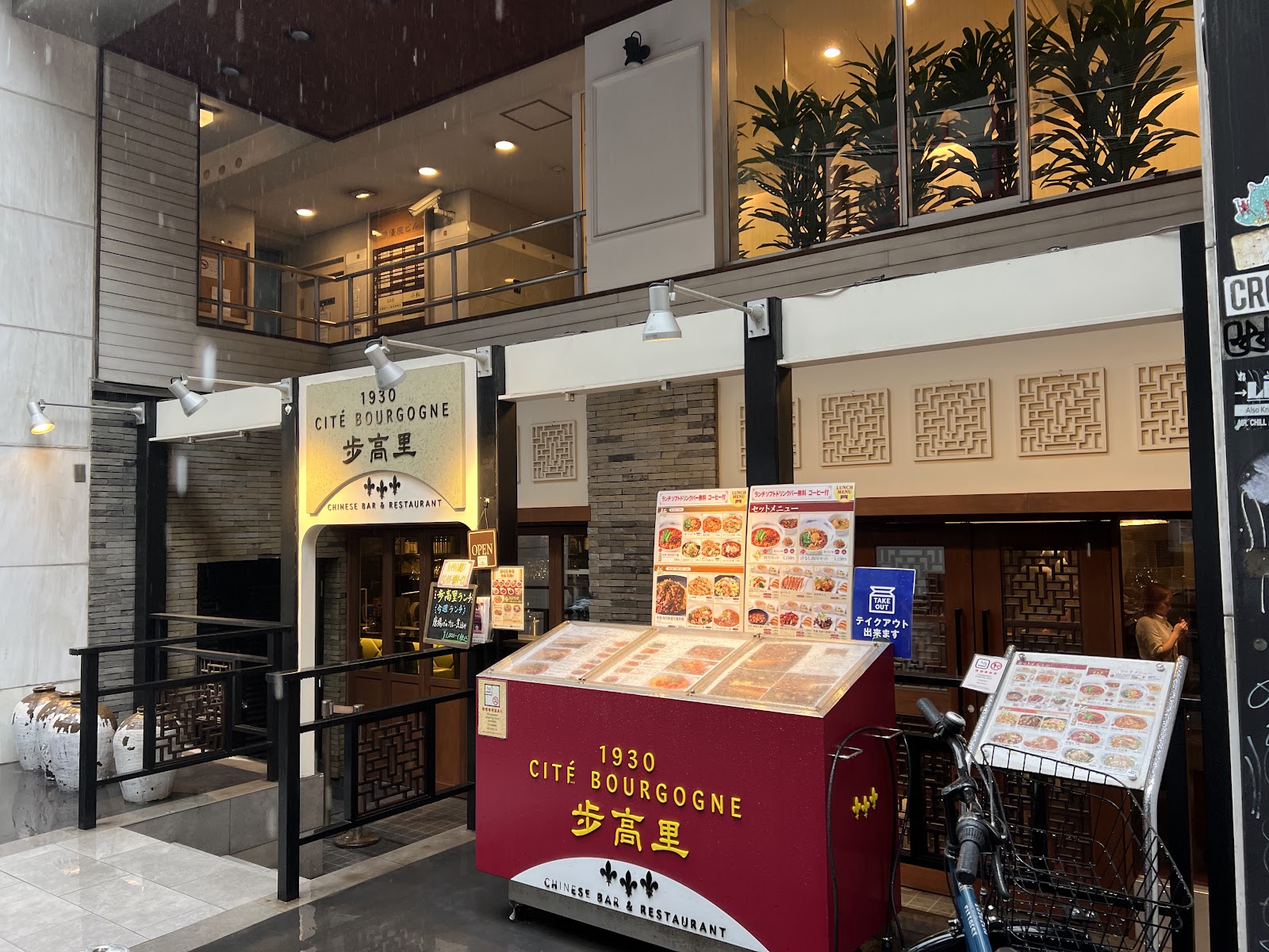 雨の日の店先。金文字の看板と赤い屋台、格子の欄干、白い甕、歩道に自転車。