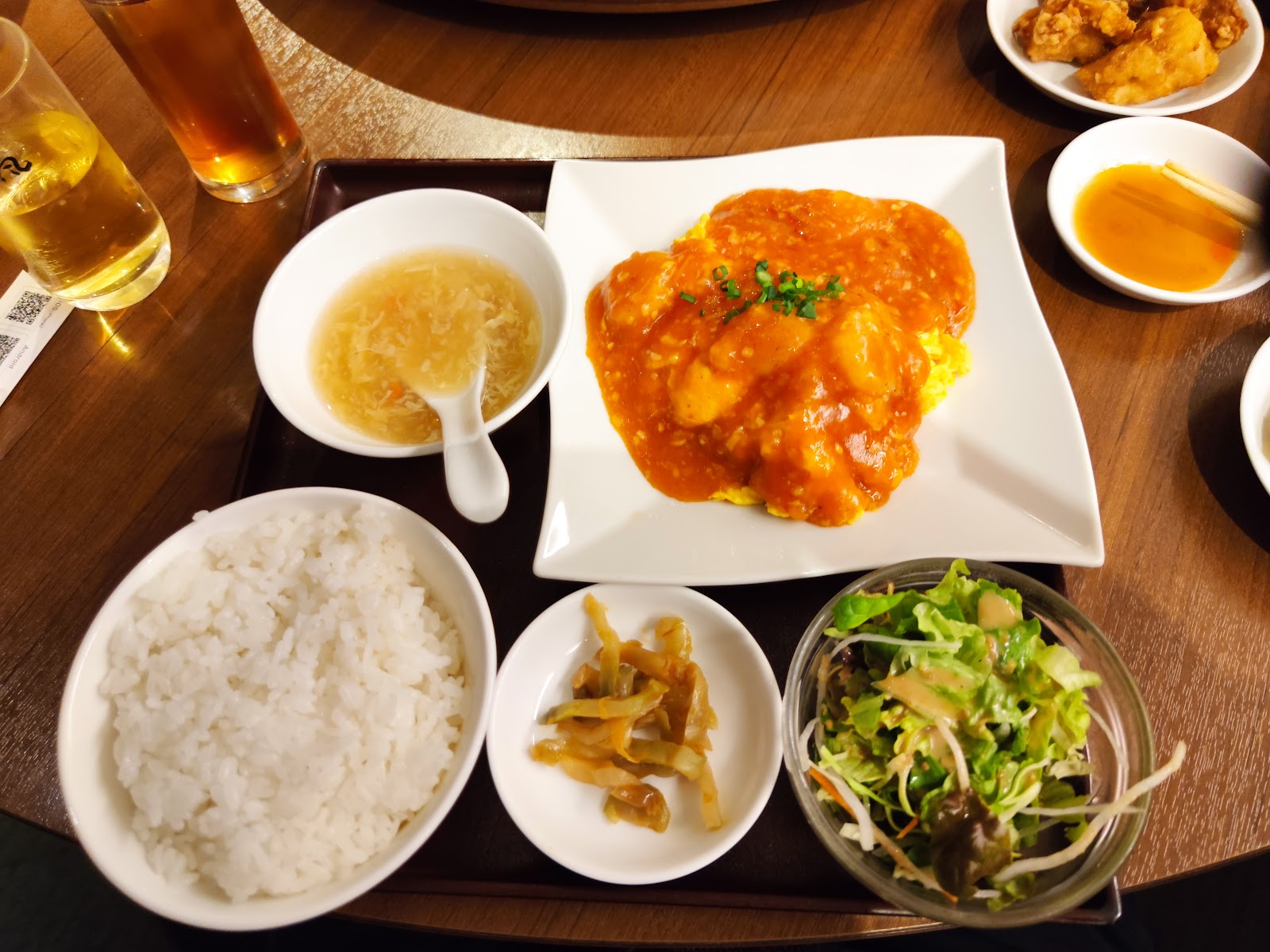 盆にのった昼の一膳。エビのチリソース炒めと卵、白いご飯、卵スープ、ザーサイ、サラダ。奥に唐揚げの小皿。